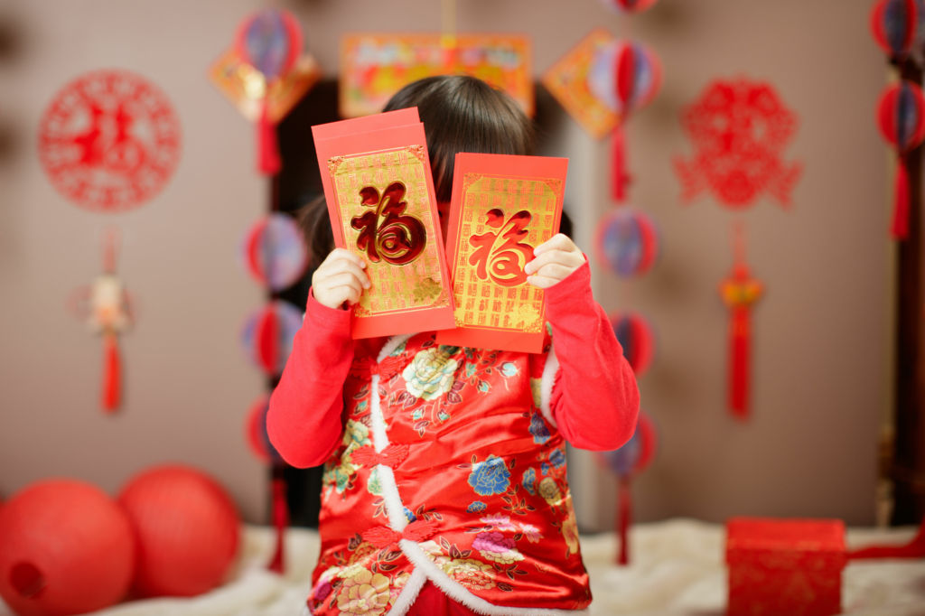 Adobe Stock - Photo d'illustration - Petite fille habillée en tenue traditionnelle du Nouvel an Chinois qui se cache avec des enveloppes rouges.
