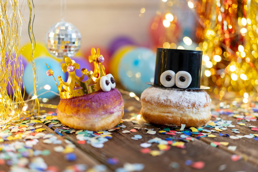 Adobe Stock - Photo d'illustration - Des beignets personnifiés, saupoudrés de sucre glace coloré qui font la fête au milieu de confettis, de rideaux scintillants et de ballons.