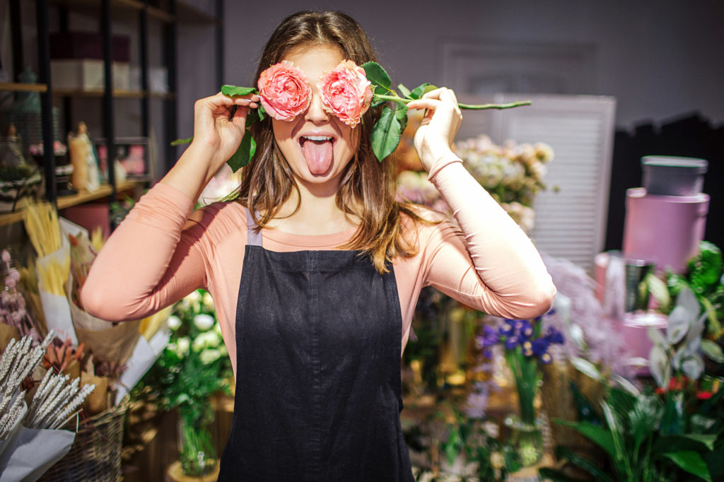 Adobe Stock - Photo d'illustration - Une jeune fleuriste avec deux fleurs devant ses yeux, tirant la langue pour faire un visage drôle.