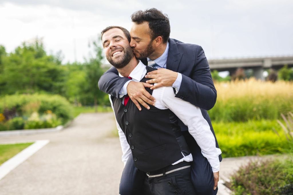 Adobe Stock - Photo d'illustration - Un homme saute sur le dos de son mari et lui embrasse la joue à la suite de leur mariage.