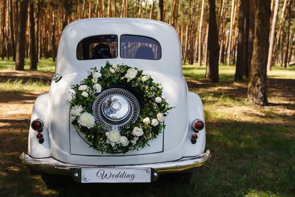Adobe Stock - Une voiture de mariage vintage de couleur blanche, style coccinelle, avec à l'arrière une couronne de fleurs. La voiture est dans une forêt.