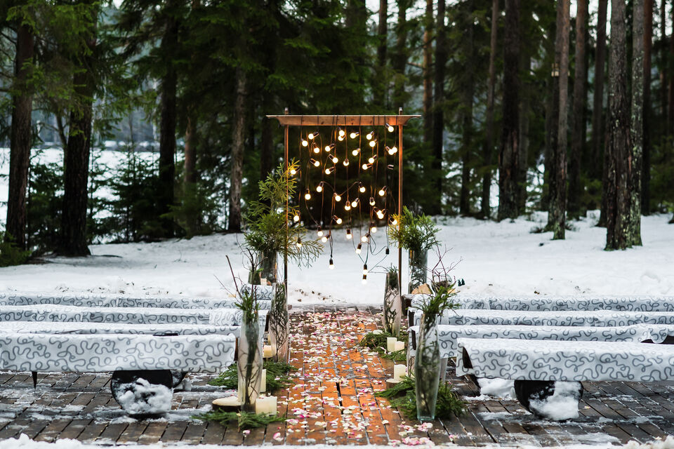 Adobe Stock - Une allée enneigée, décorée d'une arche, de plantes, de guirlandes lumineuses et de pétales de fleurs qui jonchent le sol. Une ambiance romantique propice à la saison hivernale. Les bancs sont recouverts de plaids pour protéger les invités à ce mariage du froid.<br>