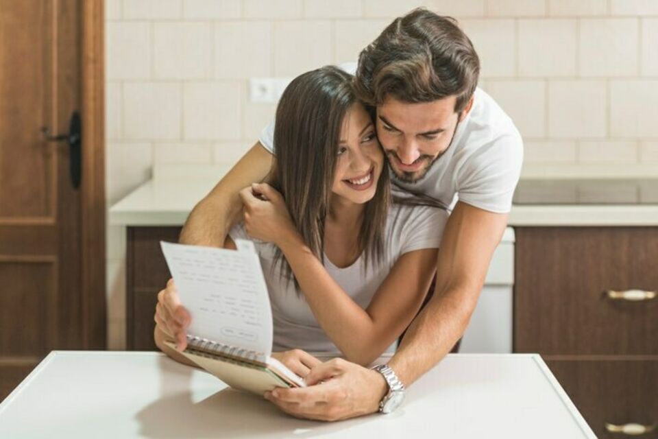 Freepik - Photo d'illustration - Une jeune femme souriante, tenant un cahier, est tendrement enlacée par son fiancé alors qu'ils planifient ensemble le timing de leur réception de mariage.
