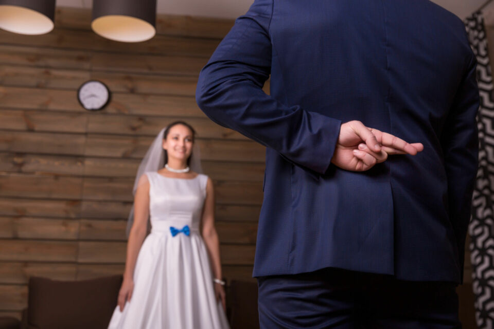 Adobe Stock - Photo d'illustration - Jeune marié croisant les doigts et redoutant la réaction de sa jeune épouse superstitieuse, lorsqu'il va lui annoncer qu'une tradition à laquelle elle tient vraiment, n'a pas été respectée.
