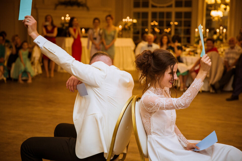 Adobe Stock -  Photo d'illustration -  Un couple joue au jeu des chaises dos à dos, levant des pancartes pour répondre à des questions amusantes sur eux-mêmes, sous les rires et applaudissements des invités.
