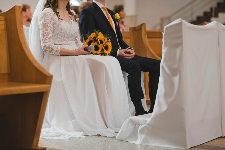 Photo d'illustration - Mariés assis côte à côte dans l’église, elle en robe blanche avec bouquet de tournesols, lui en costume sombre et cravate jaune, incarnant l’élégance et la solennité d’une cérémonie chrétienne.