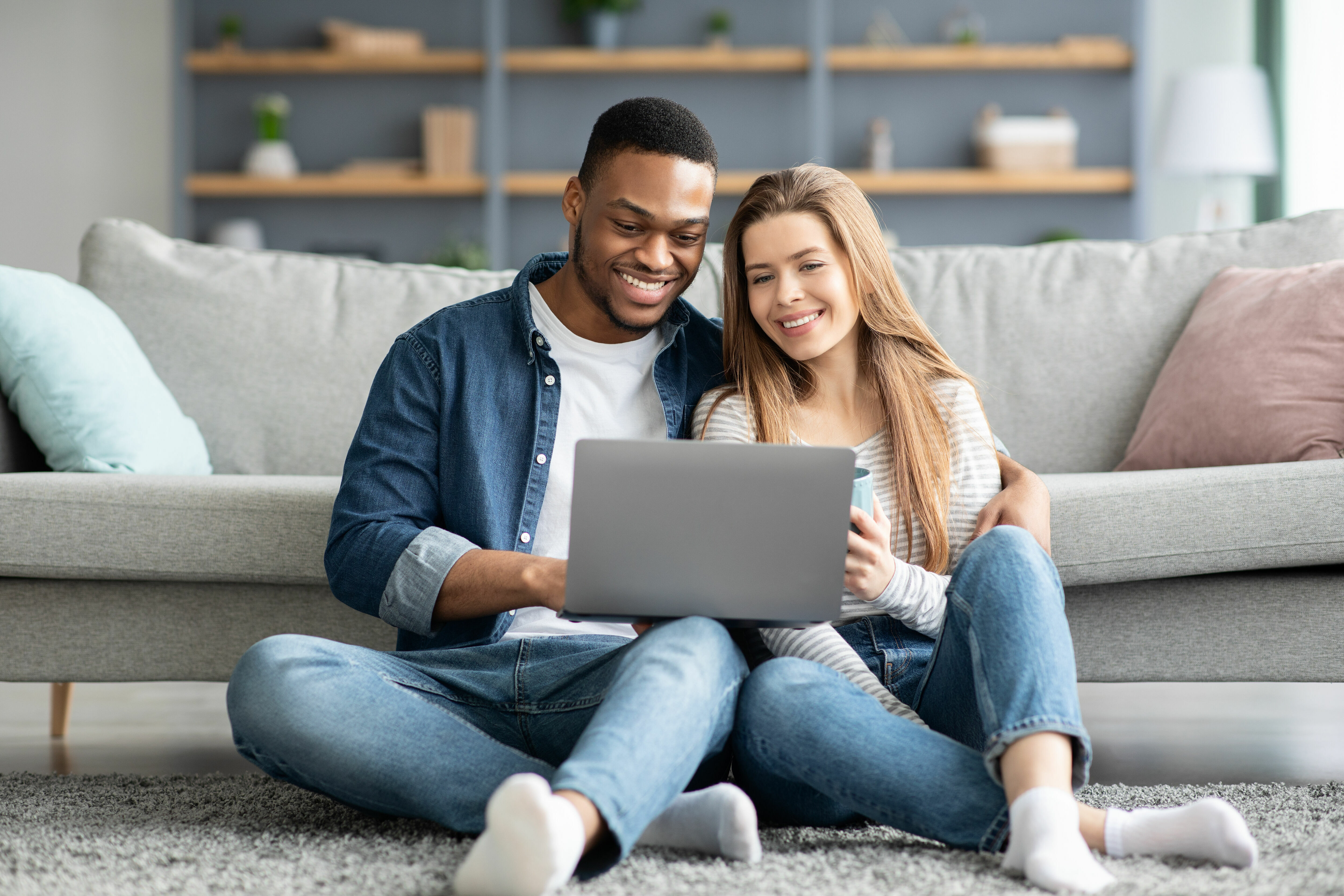 Adobe Stock - Photo d'illustration - Un jeune couple mixte, dans son salon, passant un moment devant son ordinateur portable.